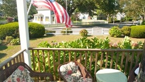 A porch with wicker furniture and a view of a house with an American flag. - Chatham Village - Adorable Studio Apartment in Antique Home (Chatham)