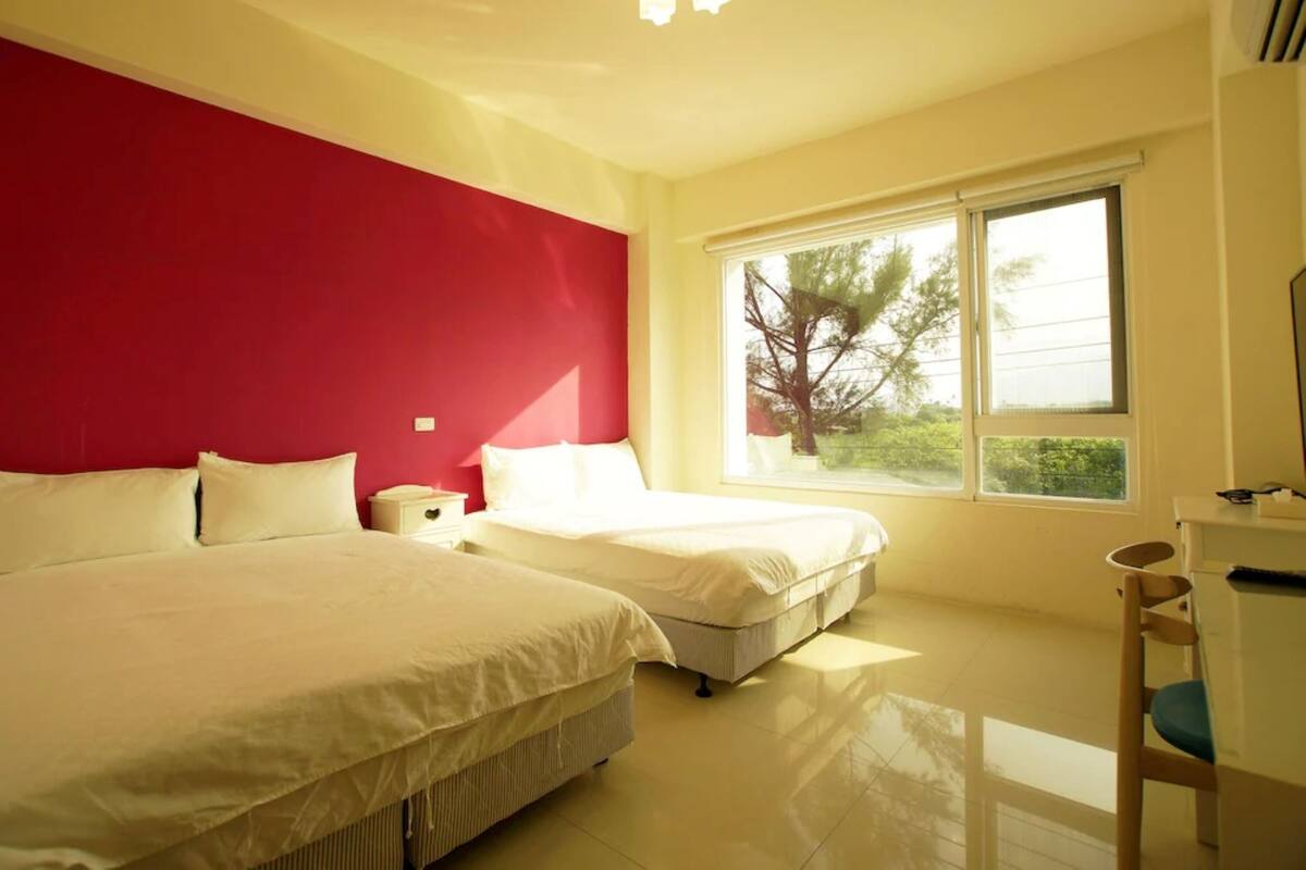 A hotel room with two beds, a red accent wall, a large window with a view, and a desk with a chair.