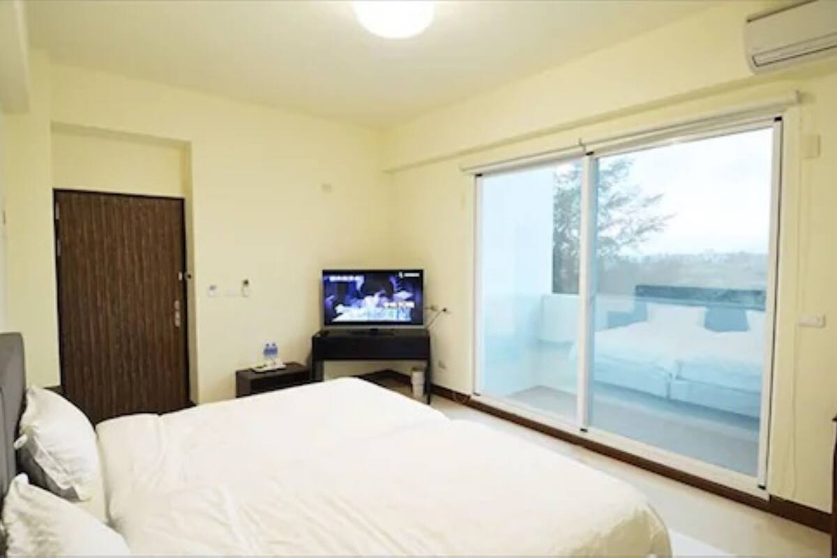 A hotel room with a bed, a TV, a sliding glass door to a balcony, and a wooden door.