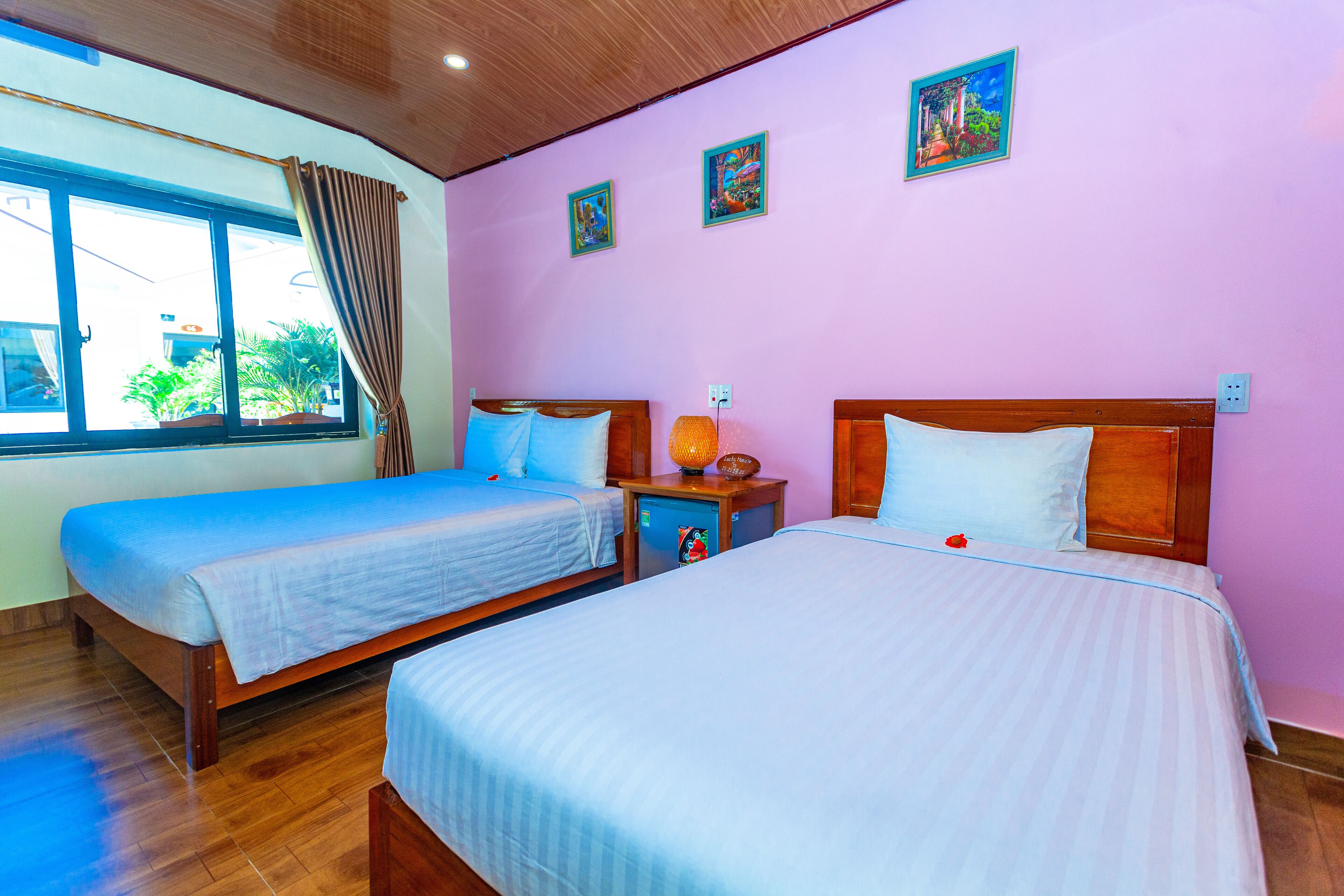 A hotel room with two beds, a window with curtains, and a wall with three small paintings.