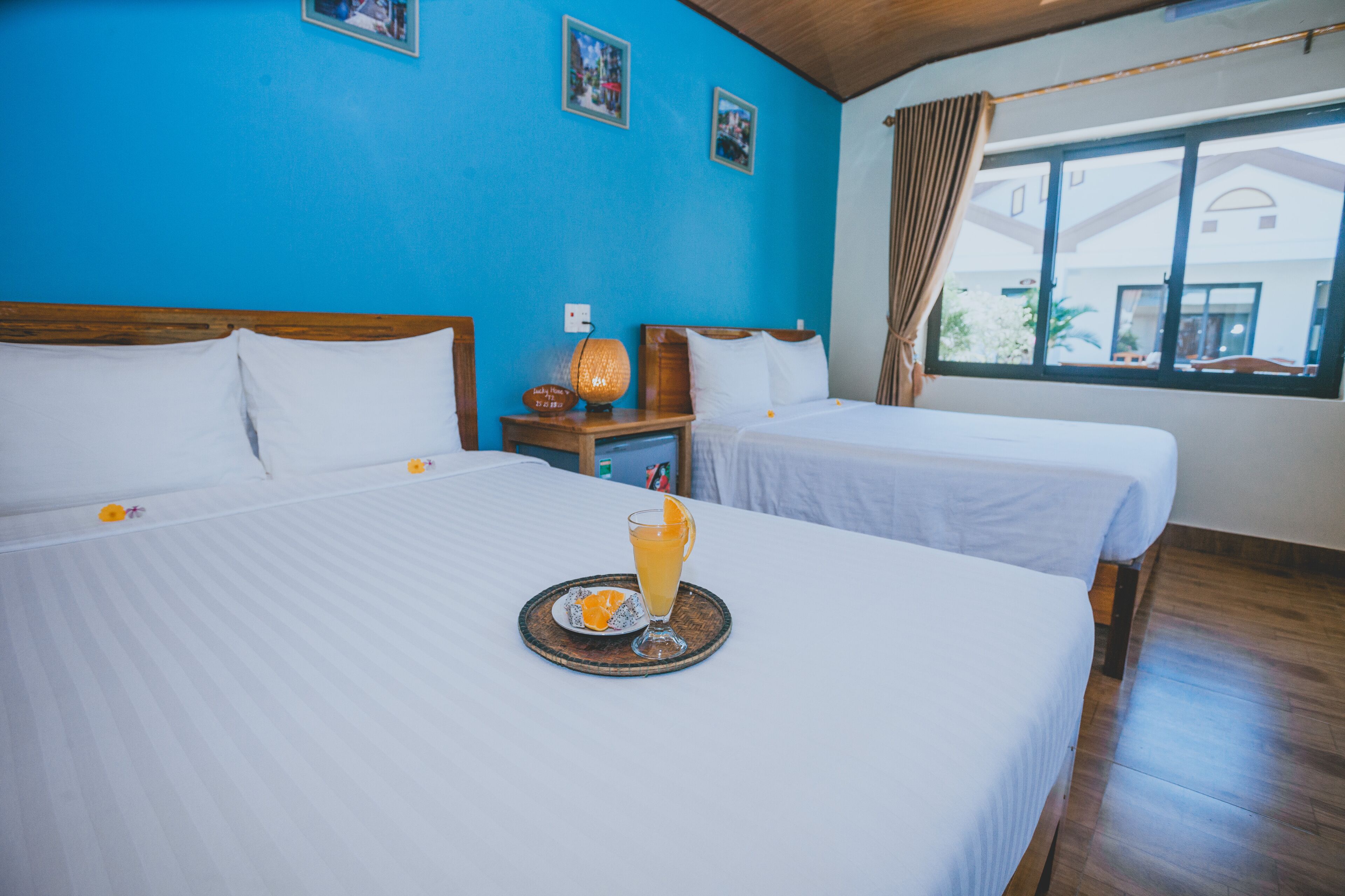 Habitación de hotel con dos camas, piso de madera, ventana con cortinas y una bandeja de desayuno con jugo de naranja.