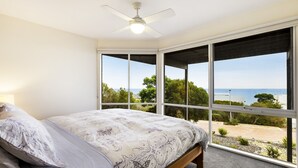 A bedroom with a bed, a ceiling fan, and large windows offering a view of the sea and greenery.