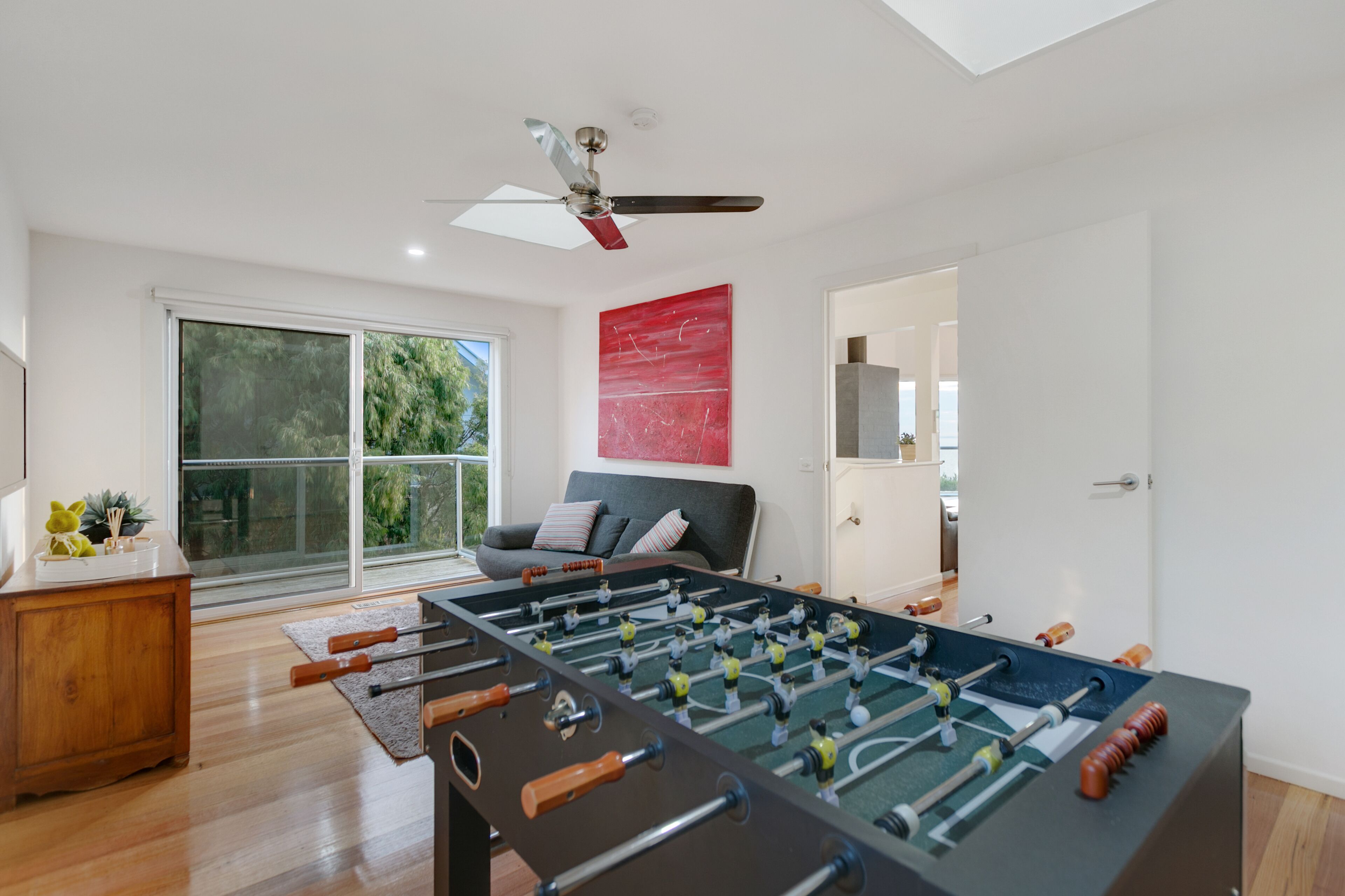 A modern living room with a foosball table, a ceiling fan, a sofa, and a large window with a view of greenery.