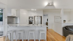 A modern kitchen with white cabinetry, a stainless steel refrigerator, and a marble countertop. There are four white bar stools and a wooden floor.