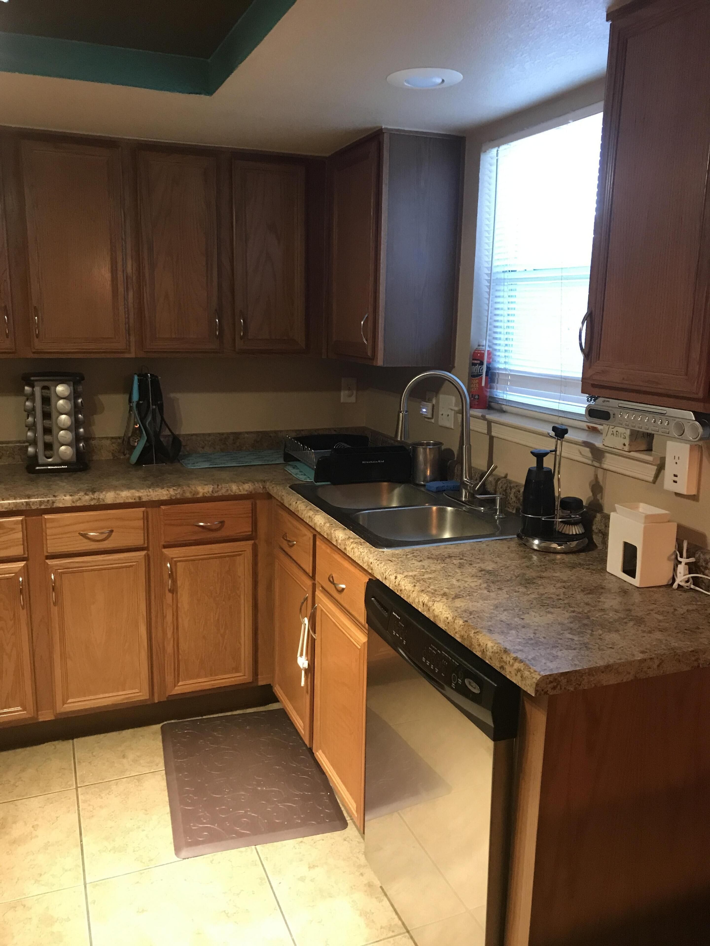 A kitchen with wooden cabinets, a granite countertop, a dishwasher, and a window with blinds.