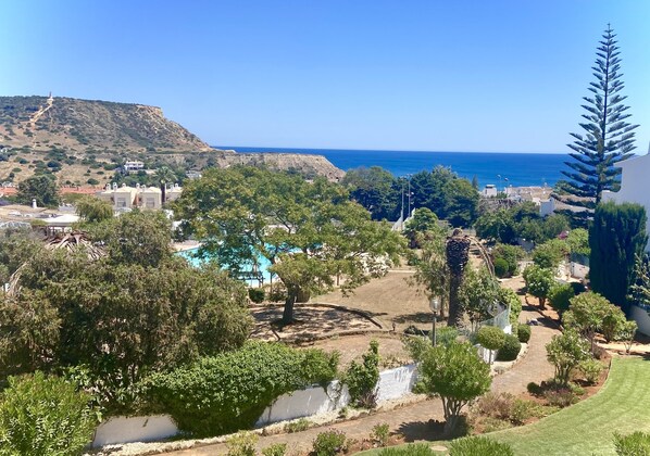 A residential area with a view of the sea, a hill with a structure on top, and a swimming pool surrounded by trees.