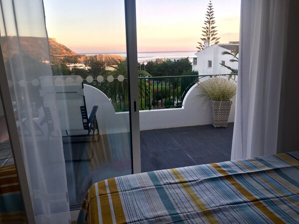 A balcony with a bed, a potted plant, and a view of the sea and buildings.