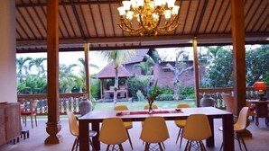 Un espace repas comprenant une table et des chaises, un lustre, et donnant sur un jardin et un bâtiment en arrière-plan.