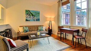 A living room with a sofa, coffee table, and chairs, a painting on the wall, and a window with a view of buildings. - Fountain View Cottage, Beautiful Savannah Home (Savannah)