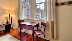 A room with a wooden table and two chairs, a lamp, and a view of buildings through large windows with curtains. - Fountain View Cottage, Beautiful Savannah Home (Savannah)