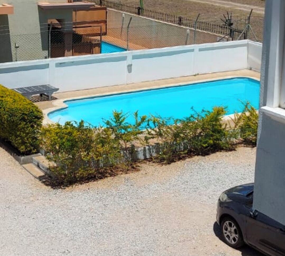 Una piscina con zona vallada, un coche aparcado cerca y un edificio al fondo.