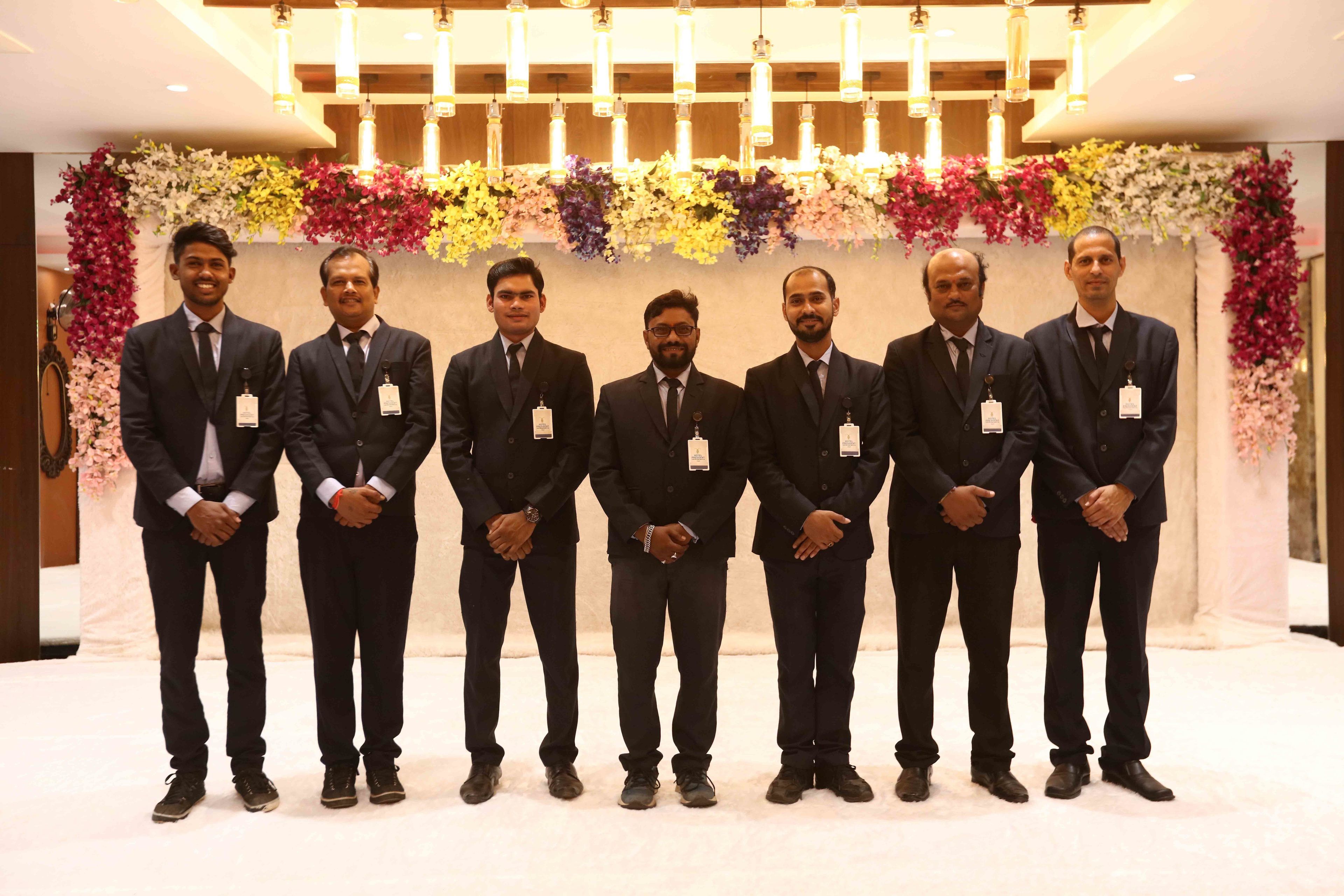 A group of men in suits standing in a room with a floral backdrop and hanging lights.