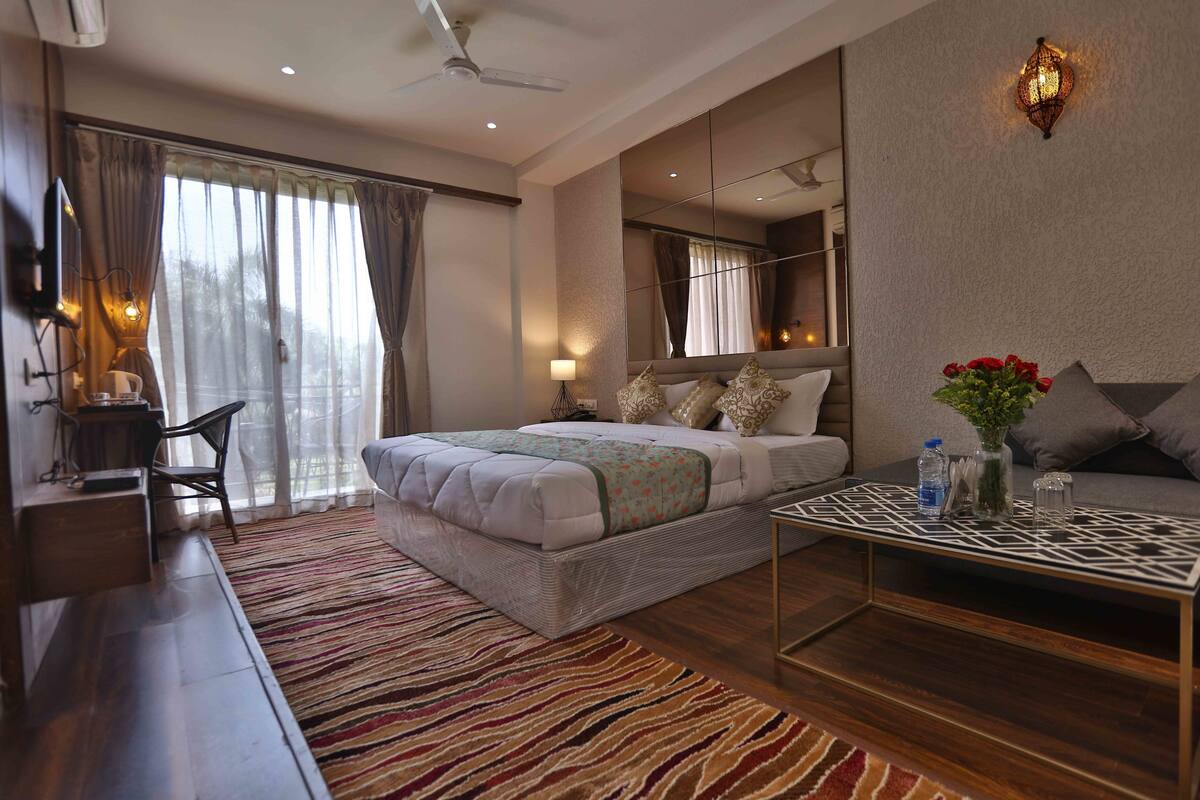 A modern hotel room with a large bed, a sitting area with a sofa, a coffee table, and a view of the outdoors through the window.
