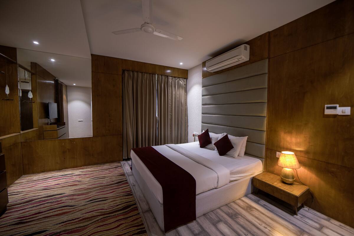 A hotel room with a bed, bedside table with a lamp, an air conditioning unit, a ceiling fan, a mirror, and a patterned rug.