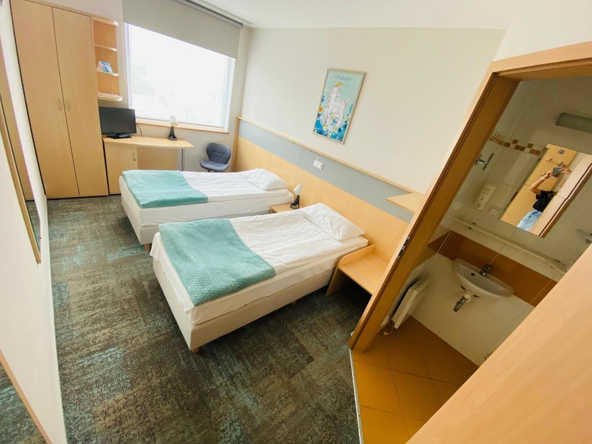 A compact hotel room with two single beds, a desk, a chair, a small table, a window, and a bathroom with a sink and mirror.