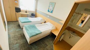 A compact hotel room with two single beds, a desk, a chair, a small table, a window, and a bathroom with a sink and mirror.
