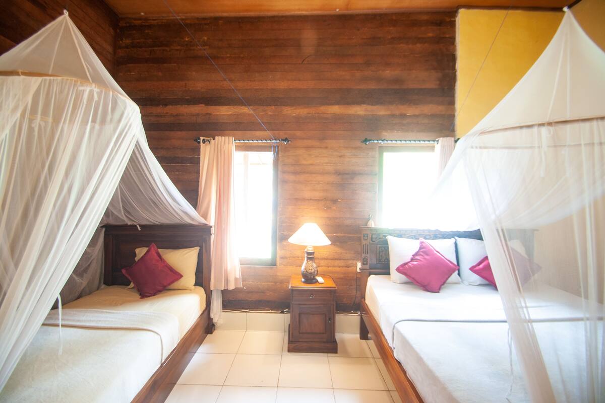 A room with two beds, each with a canopy, a small bedside table with a lamp, and a window with curtains.