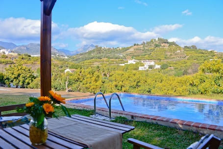 A pool with a view of a hillside, a wooden table with a vase of flowers, and a pergola structure.