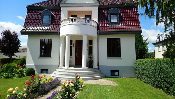A two-story house with a central balcony, surrounded by a well-maintained garden with various flowers and a lawn. - Fewo Moltke Villa - Moltke Villa (Teterow)