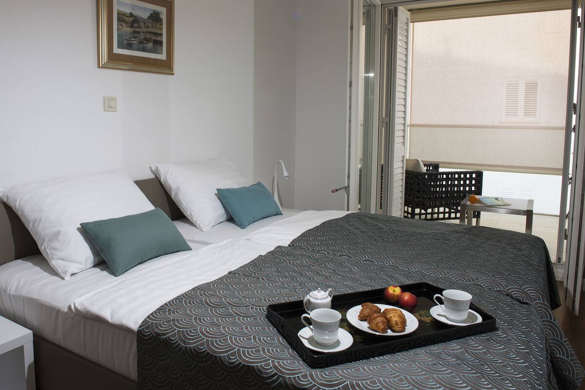 A hotel room with a bed, a bedside table, a chair, a tray with croissants, apples, and a cup.