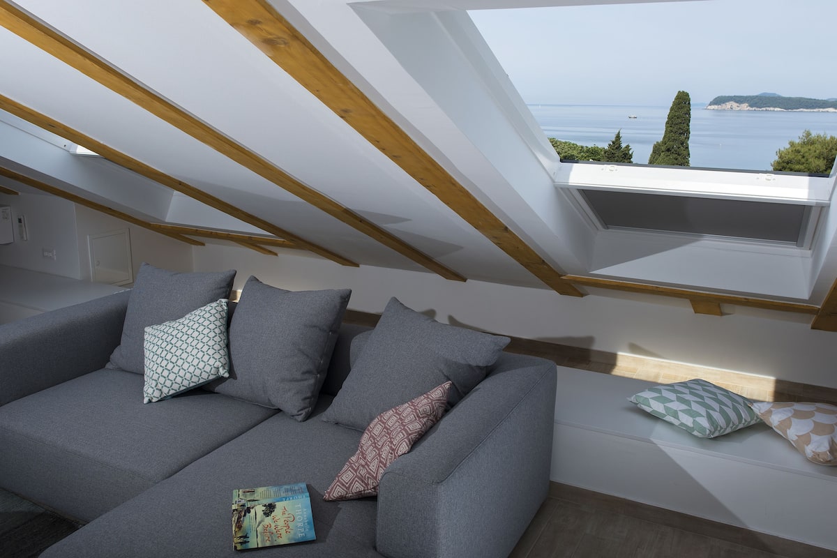 A modern loft bedroom with a sloped ceiling, a grey sofa with patterned cushions, and a view of the sea through a skylight.