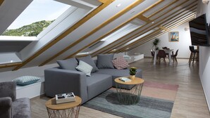 A modern loft apartment with a sloped ceiling, a grey sofa set, a dining area, and a view of a hillside. - Flowers Dubrovnik Luxury Apartments (Dubrovnik)