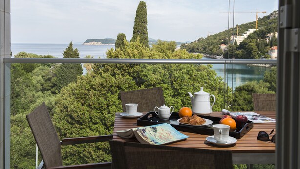 A balcony with a table set for breakfast, overlooking a coastal landscape with trees and buildings. - Flowers Dubrovnik Luxury Apartments (Dubrovnik)