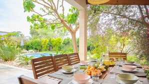 Un área de comedor al aire libre con piscina, sillas de madera y una mesa dispuesta con cuencos y vasos.