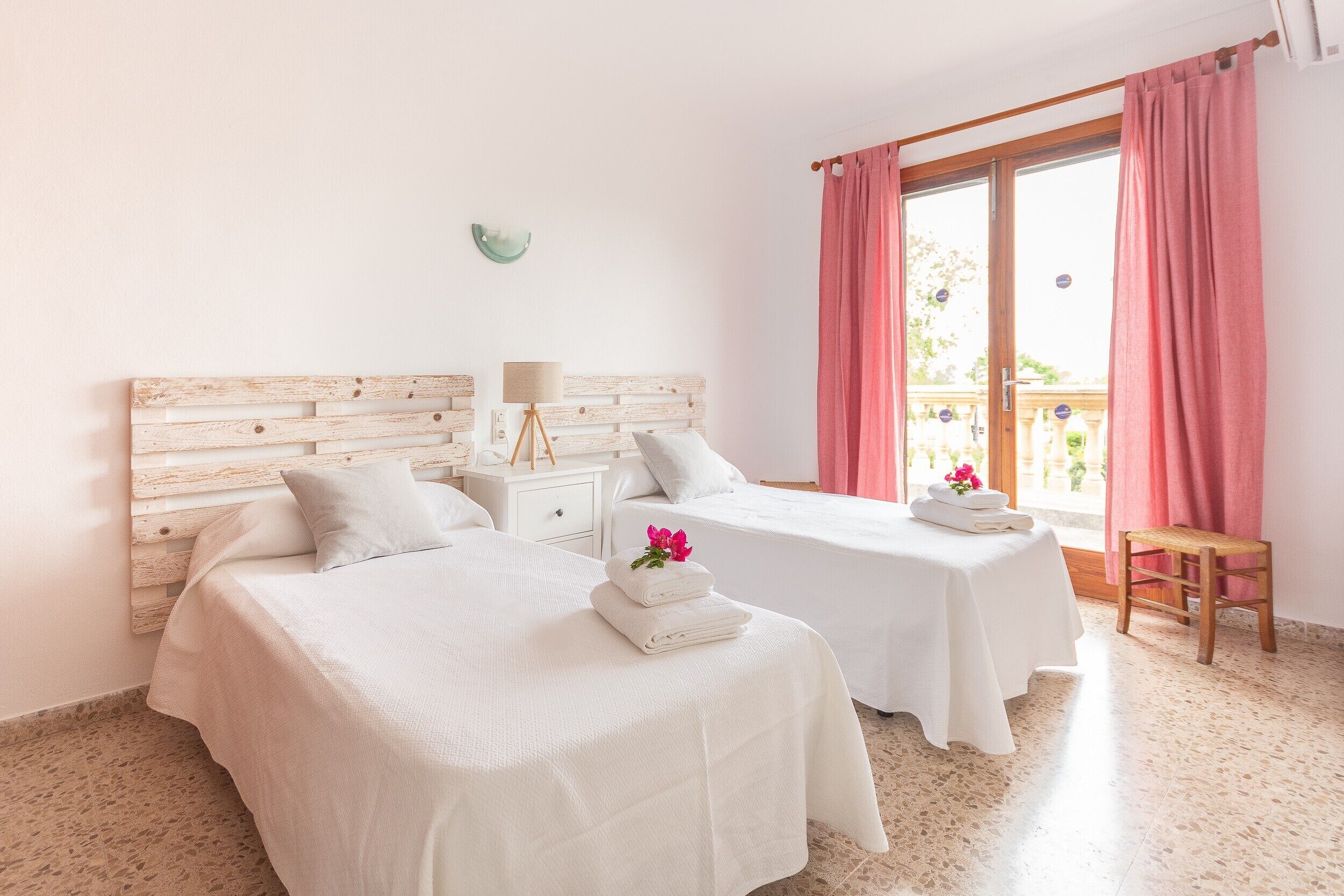 A hotel room with two beds, a small table with towels, a nightstand, and a view of the outdoors.