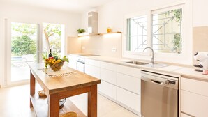 Una cocina moderna con armarios blancos, una mesa de madera con frutero, lavavajillas y ventana con vista a vegetación.