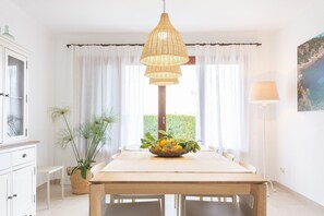 Een eetruimte met een houten tafel, een hanglamp, een potplant en uitzicht op groen.