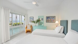 Une chambre d’hôtel avec deux lits, un balcon donnant sur la mer, un ventilateur de plafond et une petite table avec une lampe.