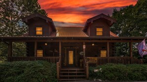 A log cabin with a porch and two attached structures, surrounded by greenery.