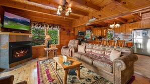 A cozy cabin interior with a fireplace, wooden furniture, a television, and a kitchen area.