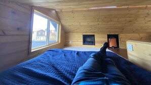 A room with a wooden sloped ceiling, a large window, and a bed with a blue quilt.