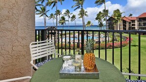 A balcony with a table set for two, overlooking a pool and resort area with palm trees and a view of the ocean.