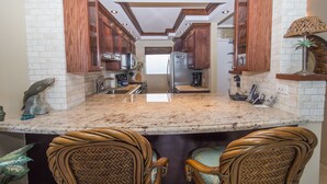 A kitchen with wooden cabinets, a granite countertop, and two wicker chairs. - Spectacular! Full A/C Penthouse Level Ocean Front 1 Bedroom - # 608 (Kihei)