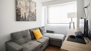 Habitació moderna amb un sofà seccional gris, un coixí groc, una taula de treball amb un ordinador i teclat, una làmpada i una finestra gran amb persianes.