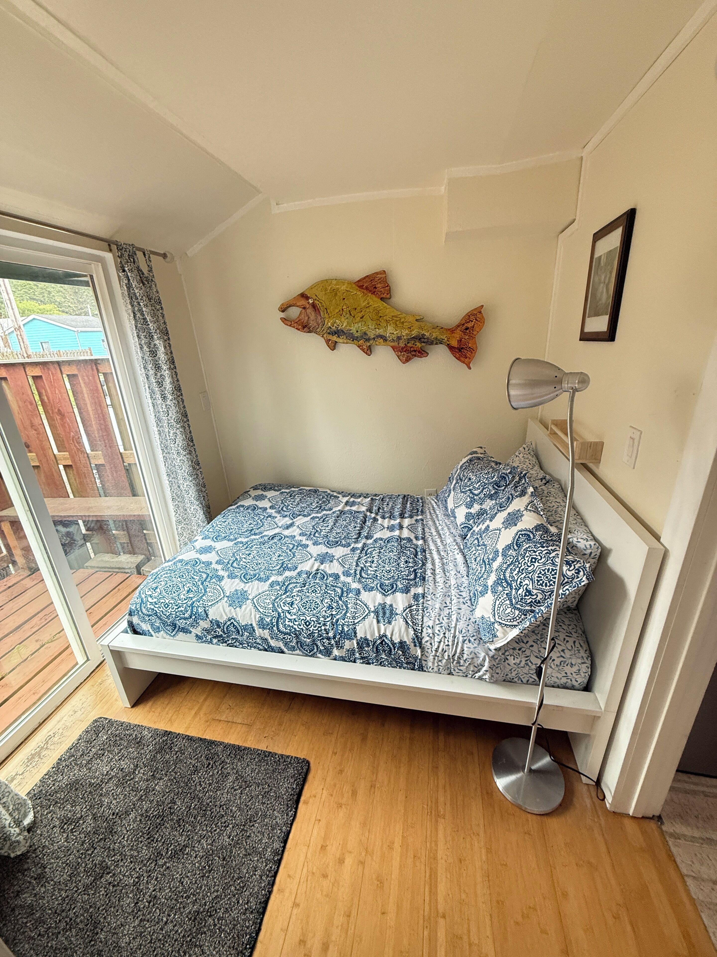 A small, single-bed room with a wooden floor, a patterned bedspread, a fish decoration on the wall, and a desk lamp.