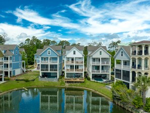 A row of multi-story houses with balconies, a small pond in the foreground, and a clear blue sky. - New Beach House, Heated Pool, Elevator, Sleeps 22 (Hilton Head Island)