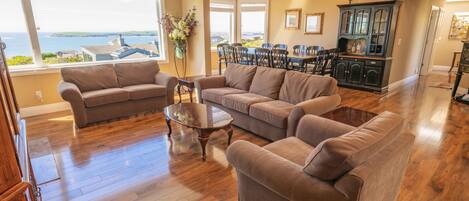 A spacious living room with hardwood flooring, a large brown sectional sofa, a glass coffee table, and a dining area with a dark wooden cabinet and chairs.