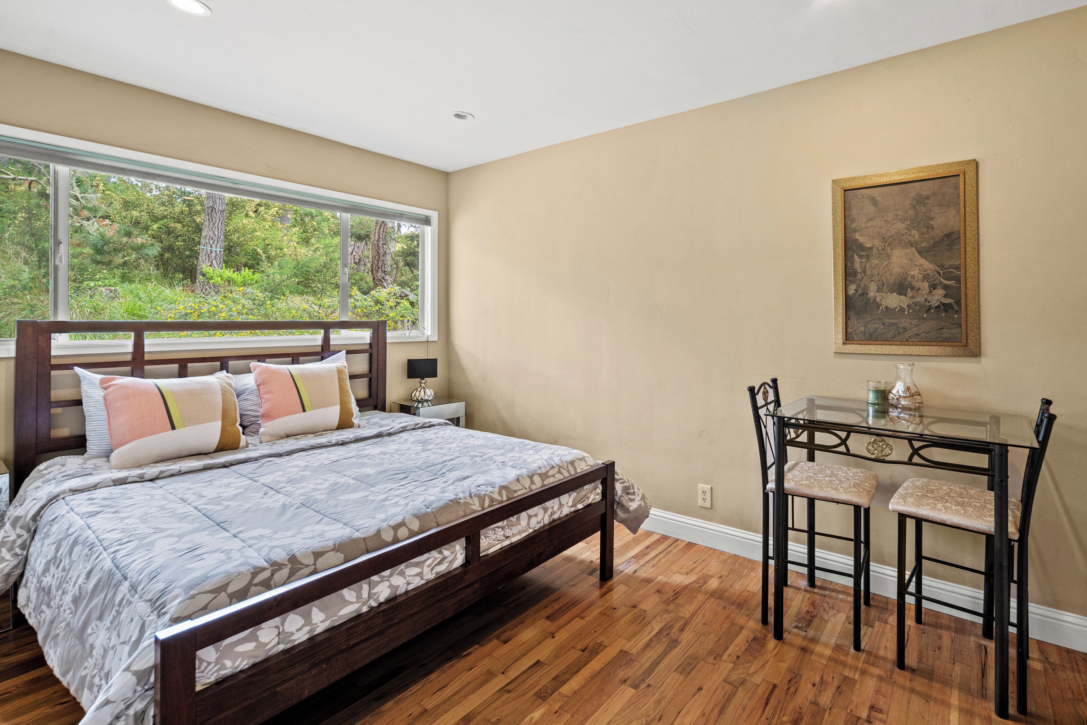 Um quarto com cama, janela, escrivaninha com cadeiras e um quadro na parede.