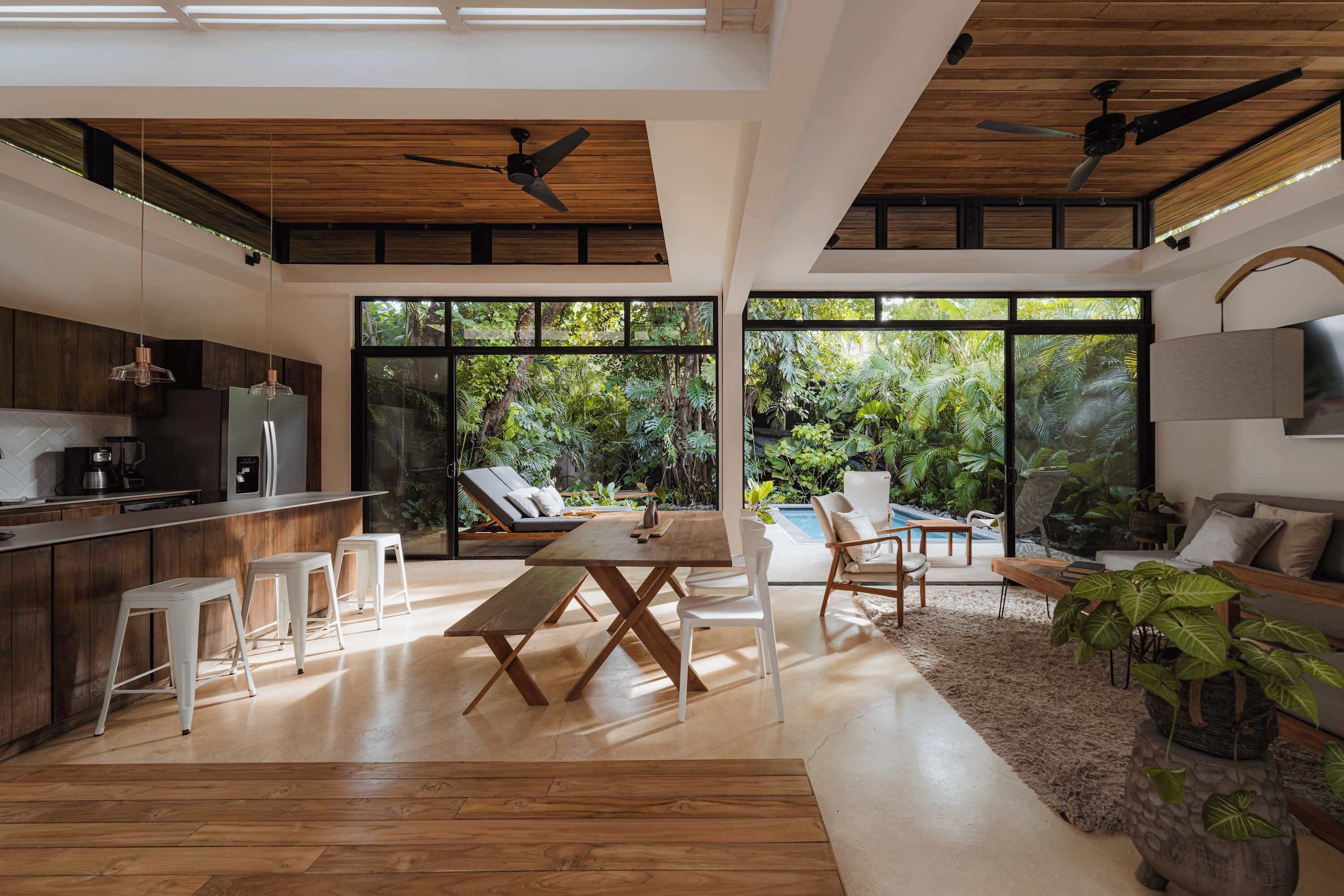 A modern kitchen with wooden cabinets, a dining area with a wooden table and white chairs, and a living area with a sofa and a TV.