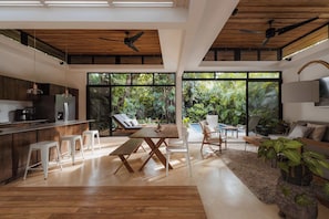 Une cuisine moderne avec des meubles en bois, un coin repas avec une table en bois et des chaises blanches, et un salon avec un canapé et un téléviseur.