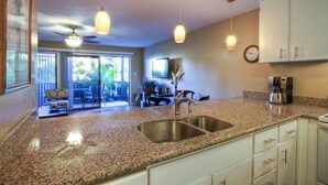 Una cocina con encimeras de granito, pileta de acero inoxidable, alacenas blancas, luces colgantes, ventilador de techo y vista al patio exterior.
