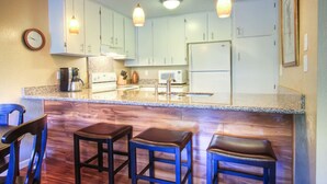 Cocina con gabinetes blancos, encimera de granito, horno empotrado, fregadero y refrigerador. Hay tres taburetes y un reloj en la pared.