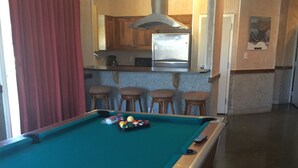 A pool table with a green felt surface, a pool cue, and a set of balls. There are wooden stools and a kitchen area with a stainless steel refrigerator and wooden cabinets in the background. - In the HEART of Ocean Drive in North Myrtle Beach (North Myrtle Beach)