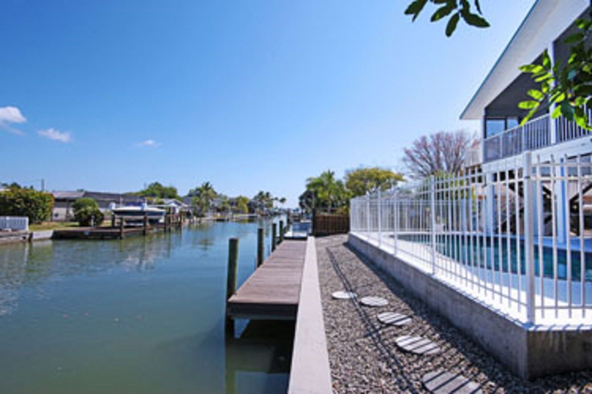 A waterfront property with a dock extending into a calm body of water, a white fence, and a building with a balcony.