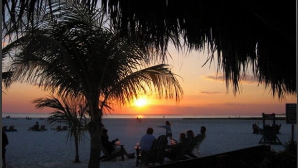 Eine Strandlandschaft bei Sonnenuntergang mit Palmen, entspannten Personen im Sand und Booten auf dem Wasser.