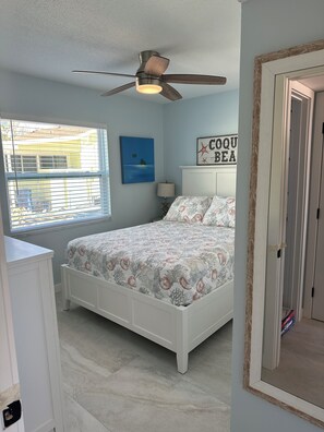 Un dormitorio con cama, ventilador de techo, ventana con persianas, un cuadro y un letrero en la pared.